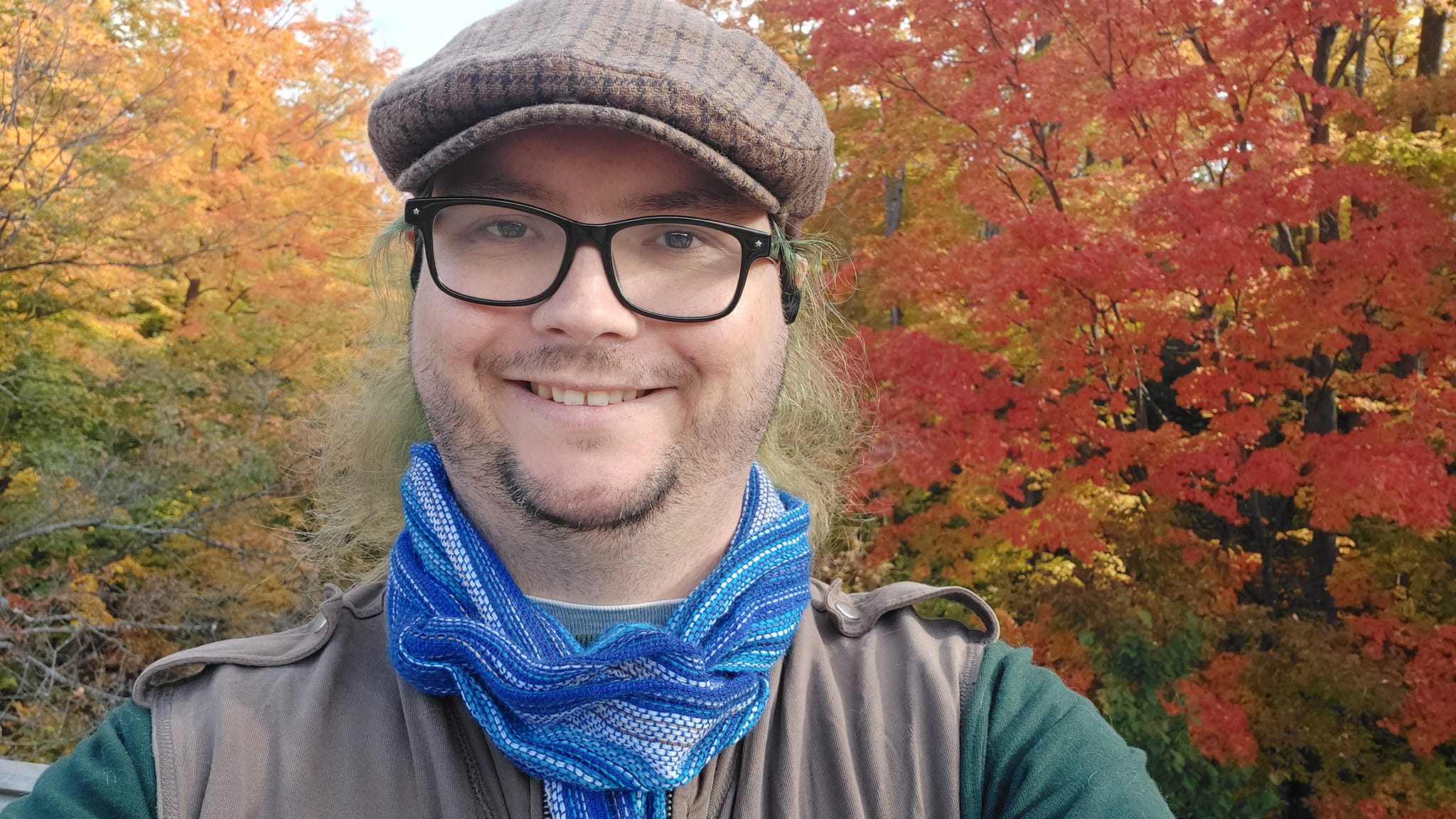 A man with long green hair wearing a green long sleave shirt with a brown zippered sweater vest, a blue scarf and a brown flat cap stand in front of a forest path with leaves all shades of orange, red and yellow